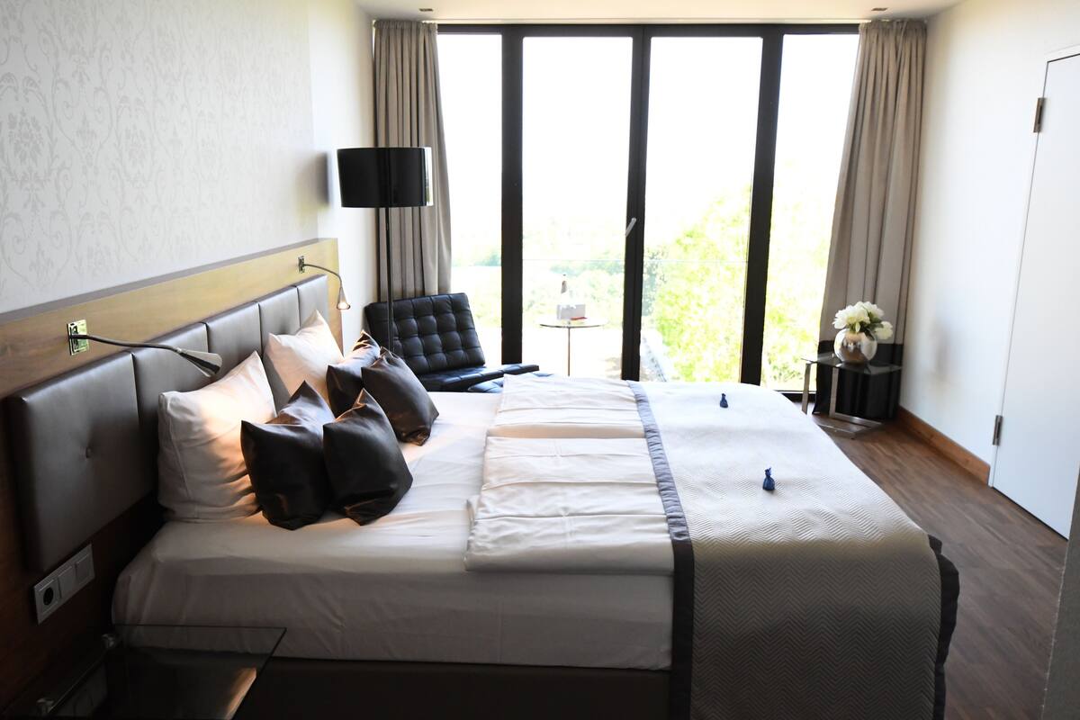 Ein Hotelzimmer mit einem großen Bett, einem Flachbildfernseher, einem Sessel, einem kleinen Tisch mit Blumenvase und Blick auf Grün hinter dem Fenster.