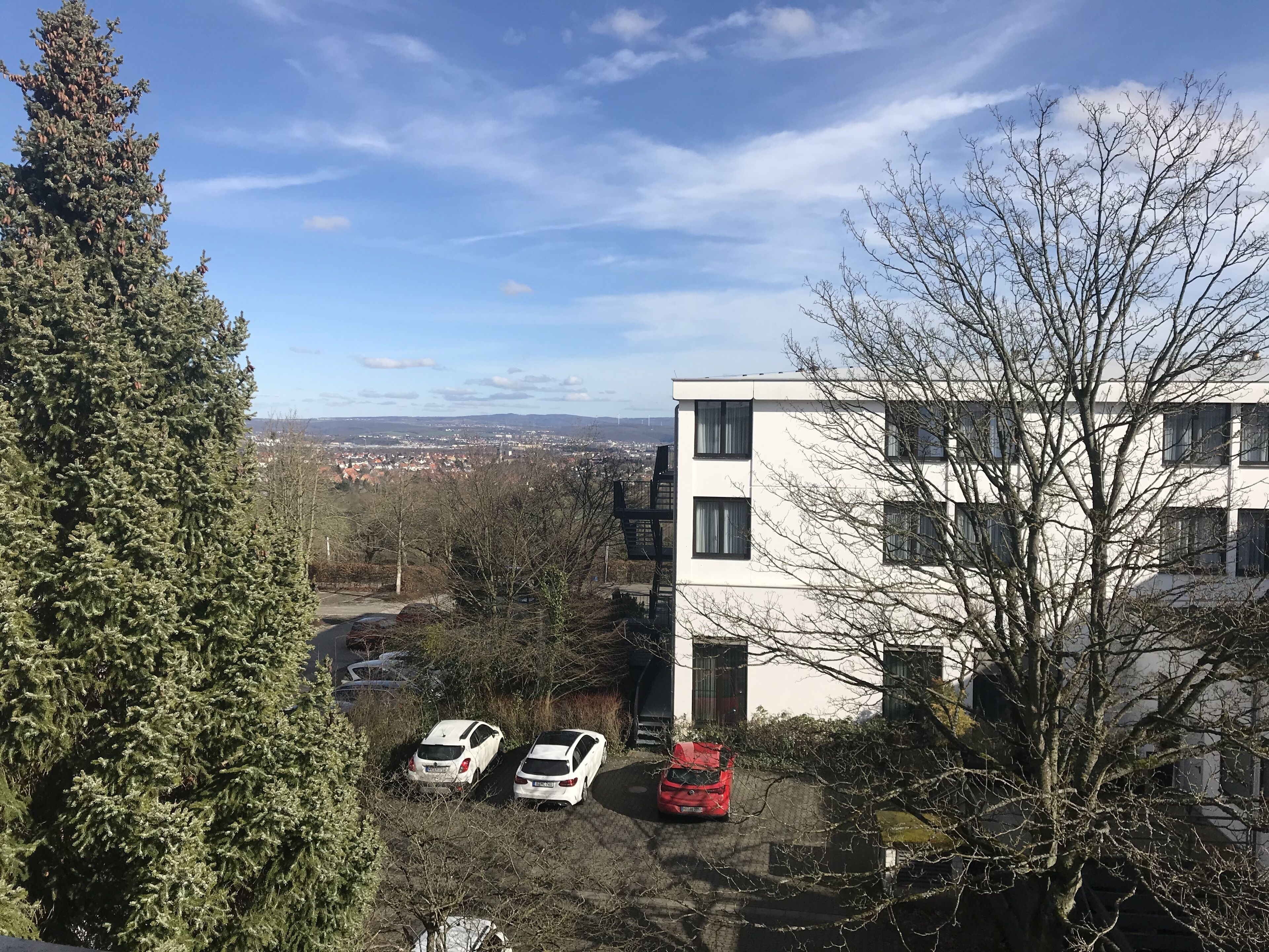 A residential area with a white building, parked cars, and a view of a distant town.