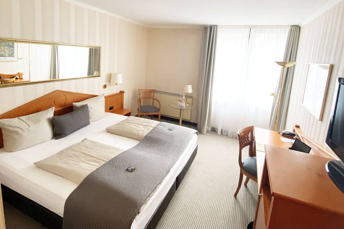 Ein Hotelzimmer mit einem großen Bett, einem Schreibtisch, einem Stuhl, einem Fernseher und einem Fenster mit Vorhängen.