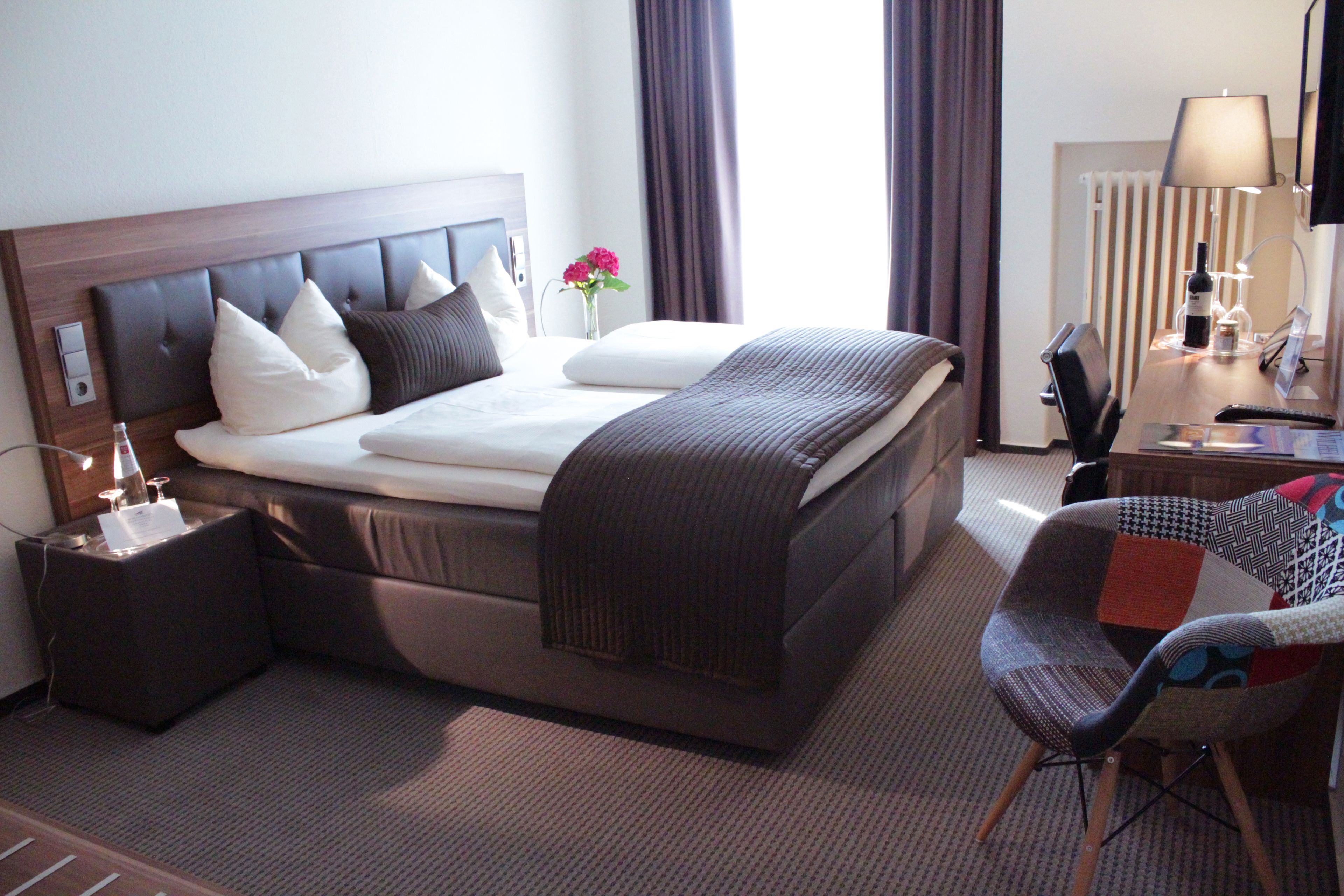 A hotel room with a large bed, a bedside table with a lamp, a chair, and a desk with a lamp.