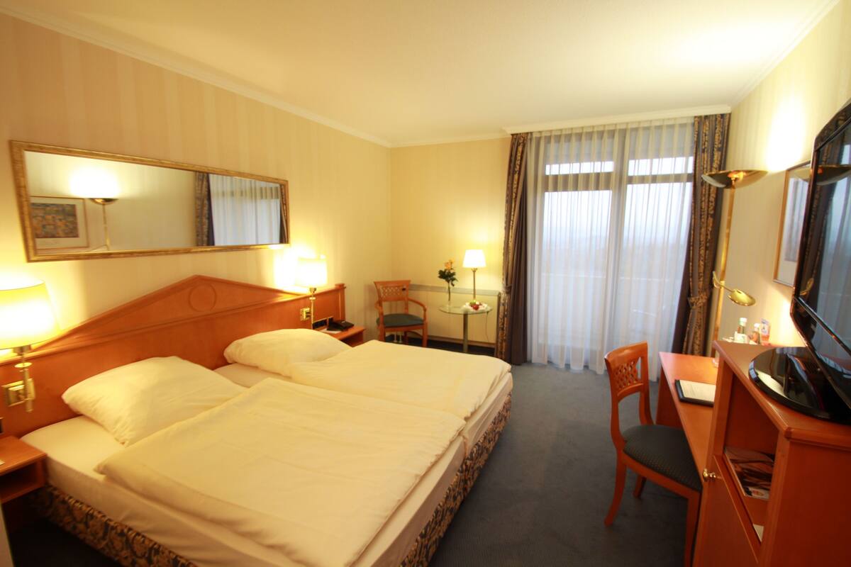 Ein Hotelzimmer mit Doppelbett, Schreibtisch mit Stuhl, Fernseher und einem kleinen Tisch mit Lampe.