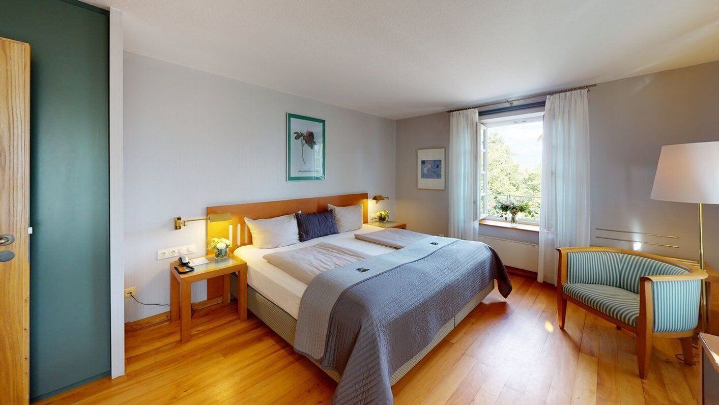 Ein Schlafzimmer mit einem großen Bett, Nachttischen, einem Sessel, einem Fenster mit Vorhängen und einem an der Wand hängenden Bild.