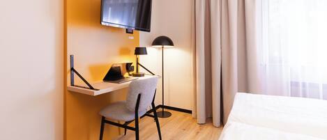 A hotel room with a bed, a desk with a chair, a TV mounted on the wall, and a lamp.