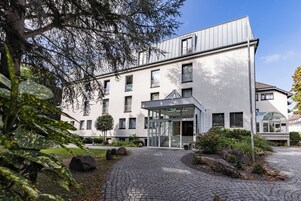 Um edifício moderno com entrada de vidro, caminho de paralelepípedos e vegetação ao redor.