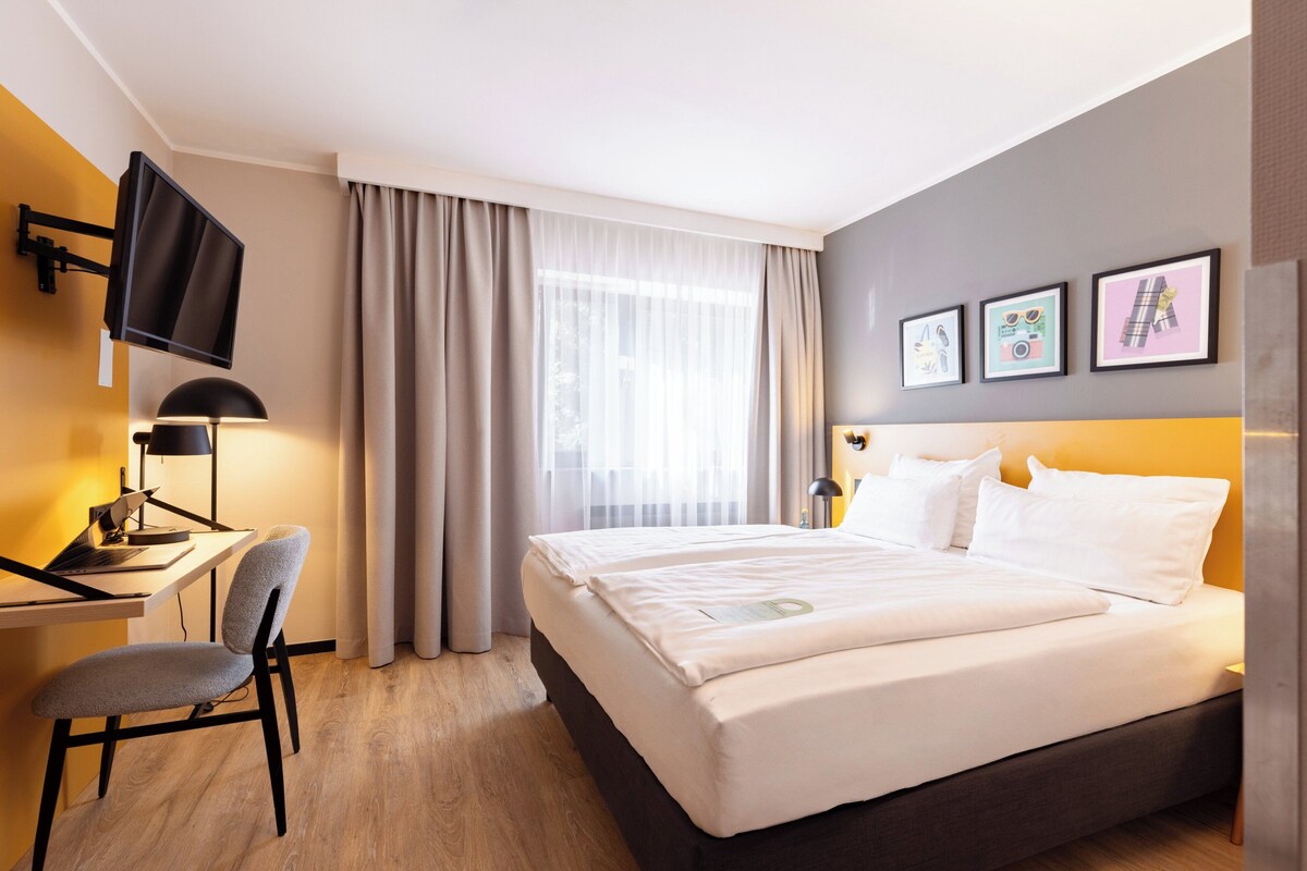 A hotel room with a bed, a desk with a chair, a TV mounted on the wall, and three framed pictures on the wall.