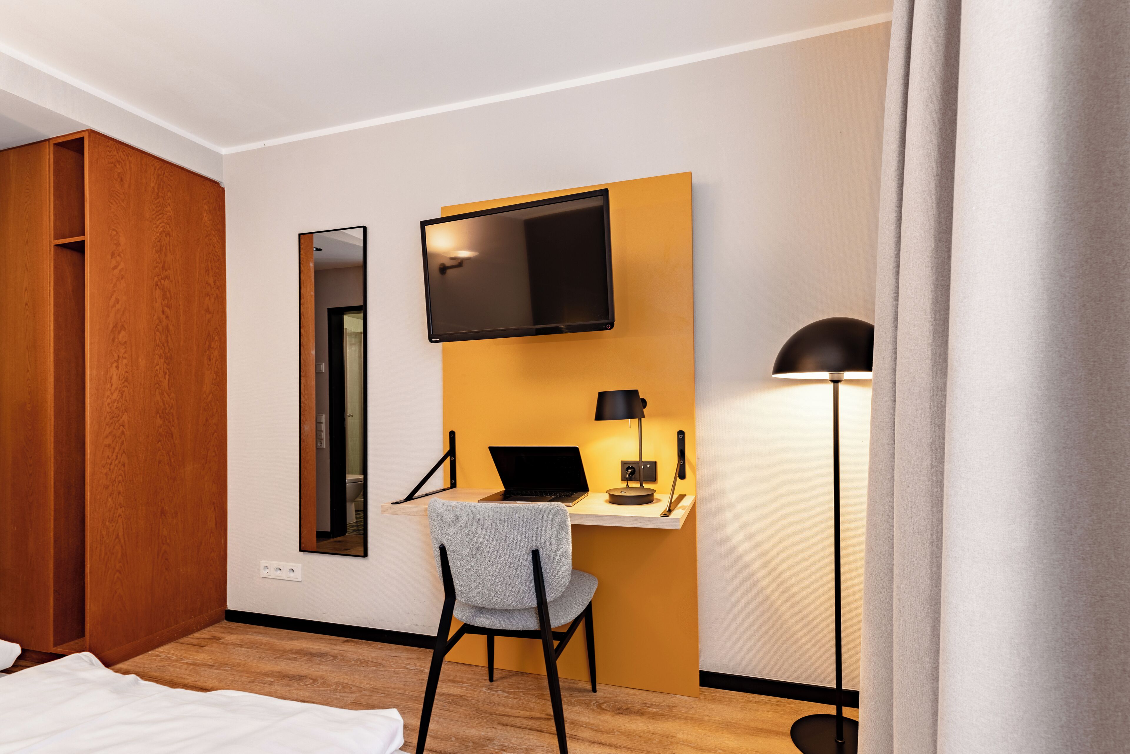 Habitación de hotel con un armario de madera, un escritorio con una computadora portátil, un televisor de pantalla plana en la pared y una lámpara de pie.