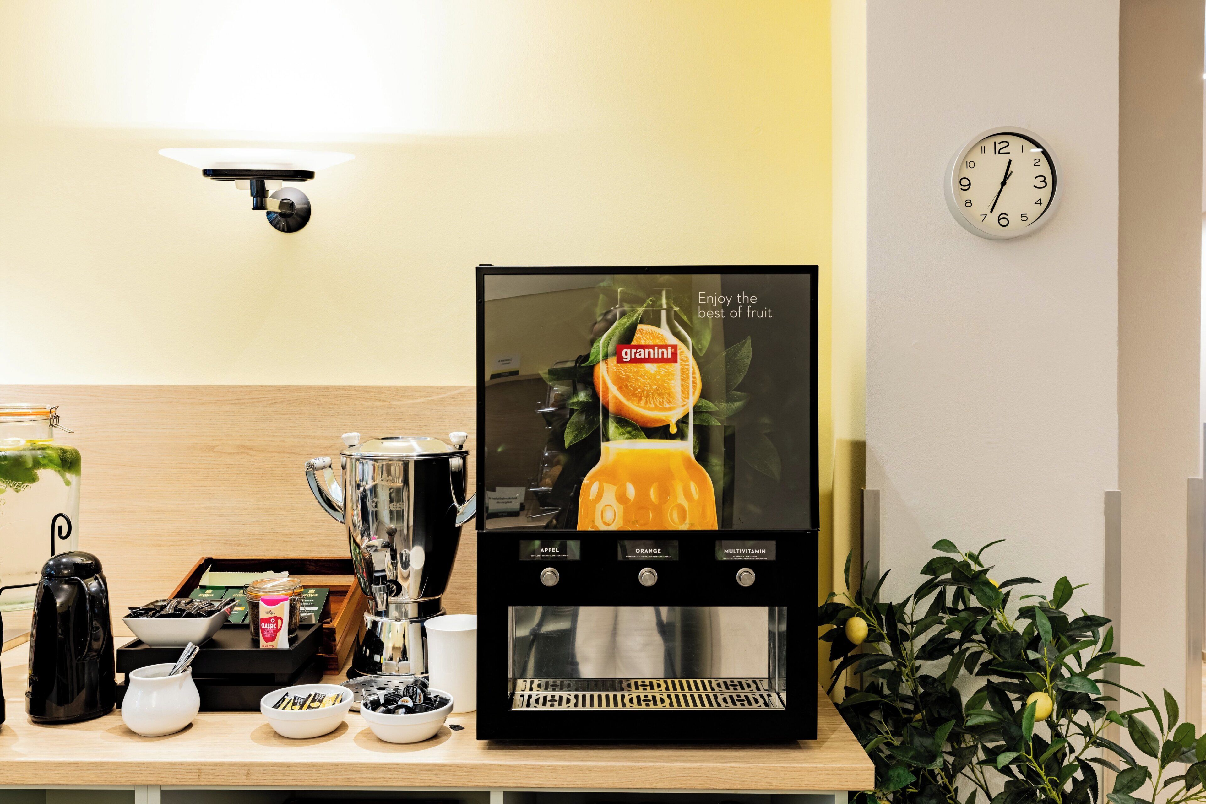 A coffee machine with a screen displaying a granini orange juice advertisement, a clock, and a tray with coffee-making supplies.