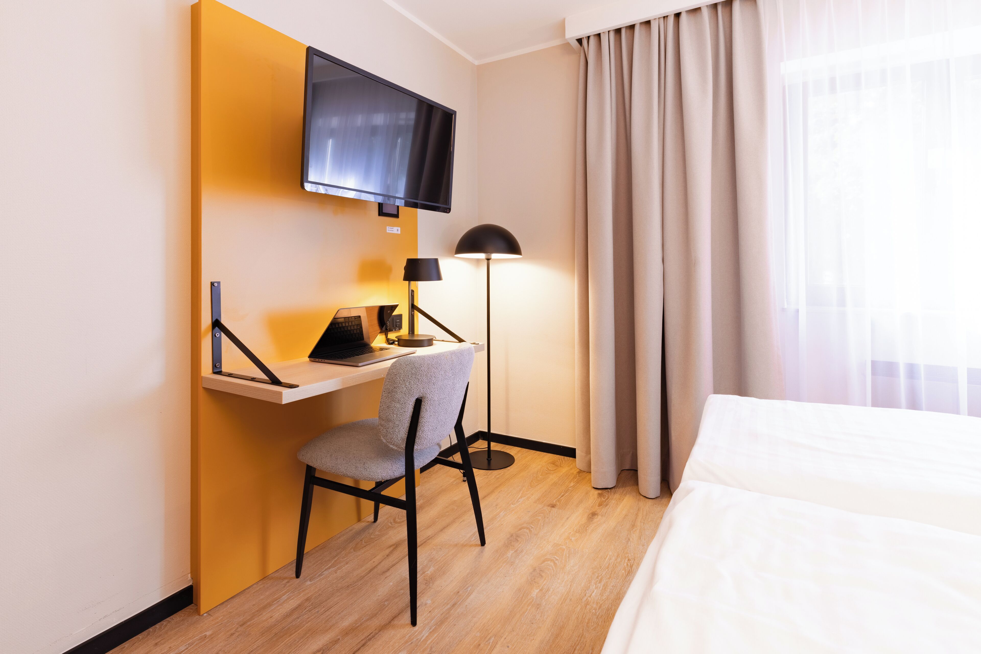 A hotel room with a bed, a desk with a chair, a TV mounted on the wall, and a lamp.