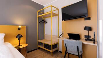 A hotel room with a bed, a desk with a laptop, a TV mounted on the wall, and a hanging rack.