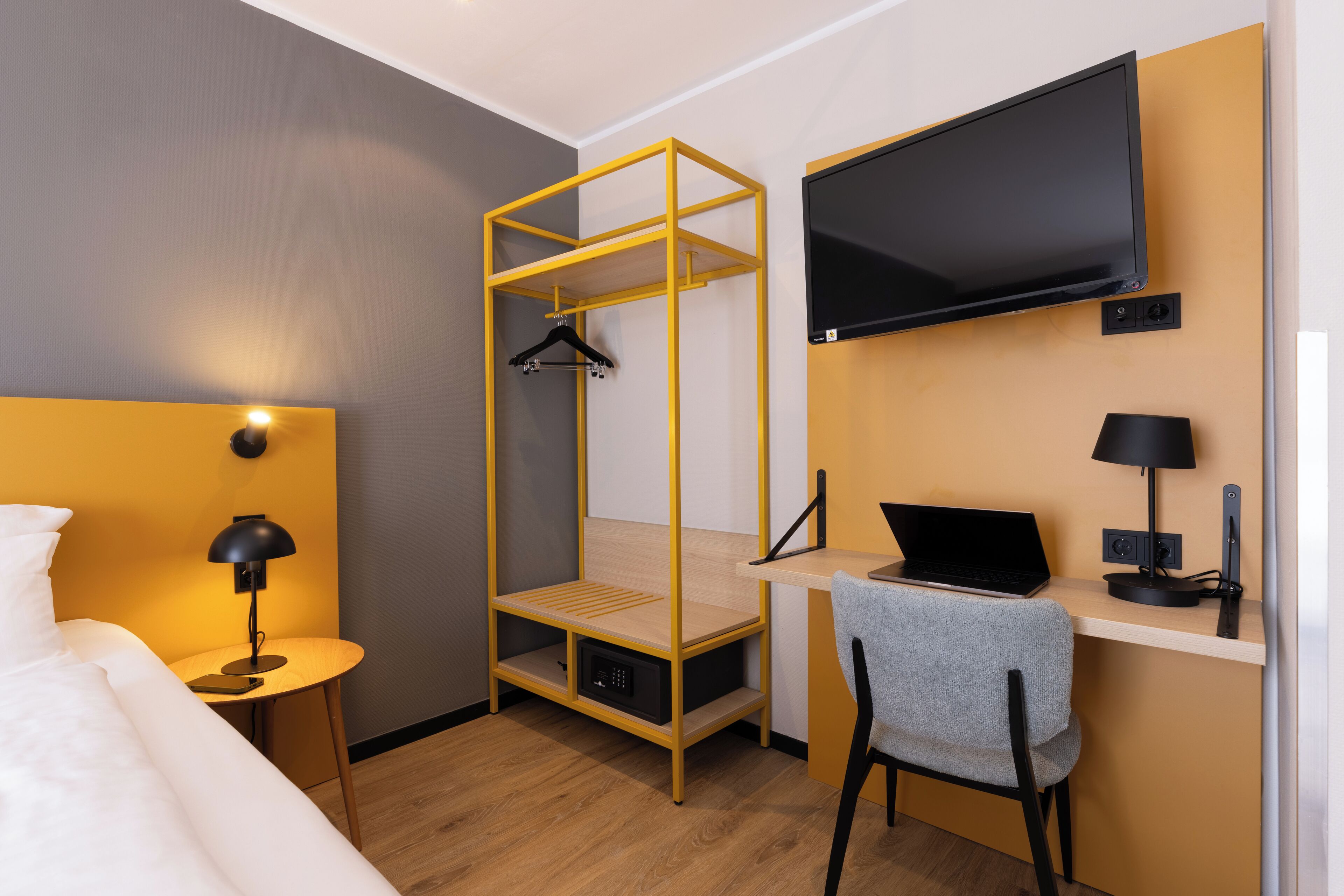 A compact hotel room with a bed, a desk with a laptop, a TV mounted on the wall, and a hanging rack.