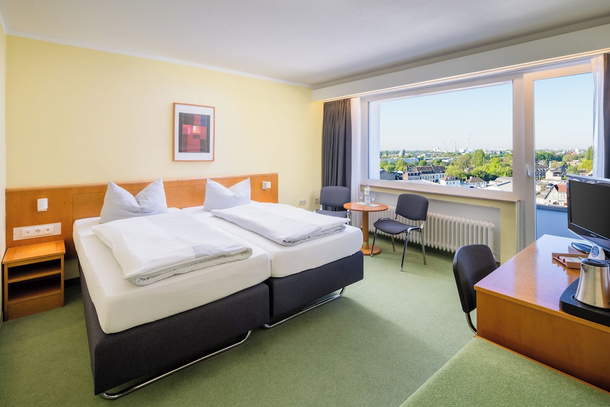 Habitación de hotel con cama doble, escritorio con computadora, una silla y vistas de la ciudad a través de amplias ventanas.