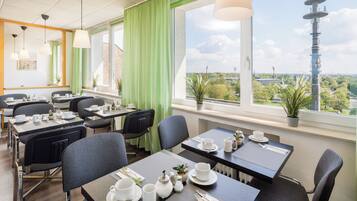 Een moderne eetruimte met gedekte tafels voor het ontbijt, grote ramen met uitzicht op een toren en groen.