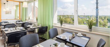 Un comedor moderno con mesas dispuestas para el desayuno, ventanas amplias con vistas a una torre y vegetación.
