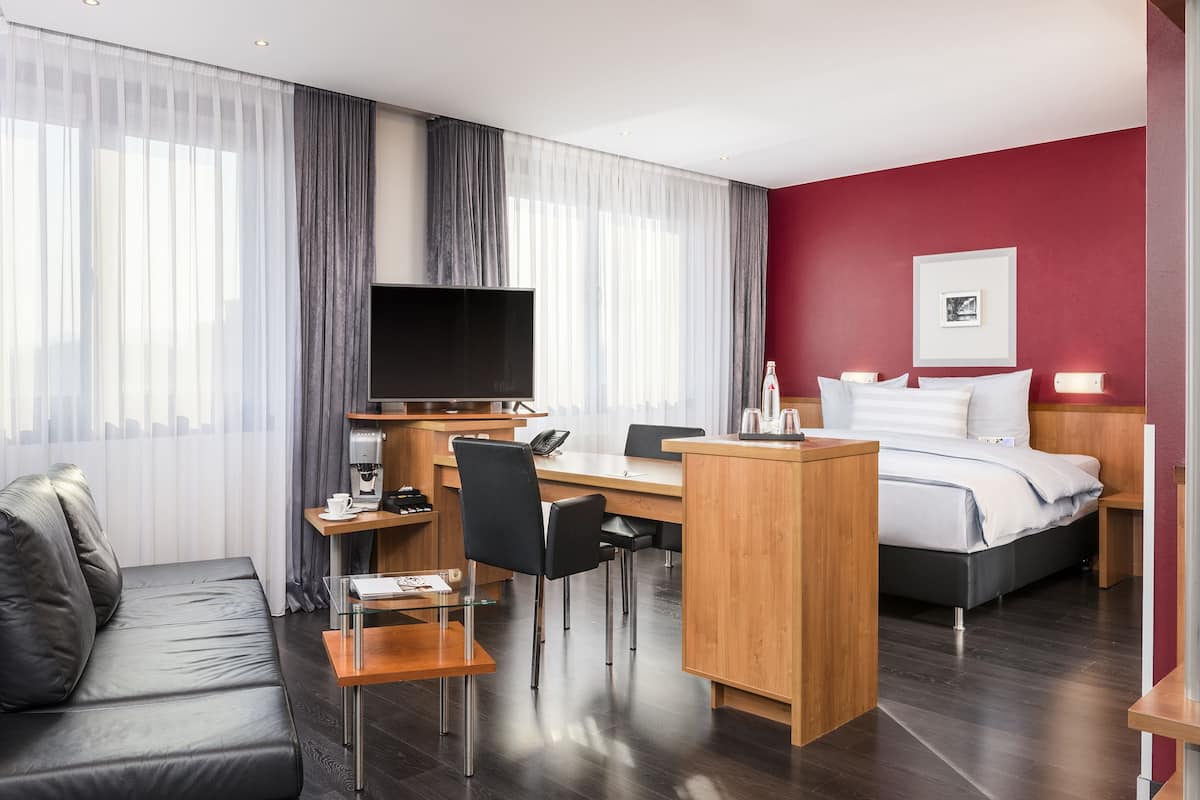 A hotel room with a bed, a desk, a chair, a sofa, a TV, and a coffee machine.