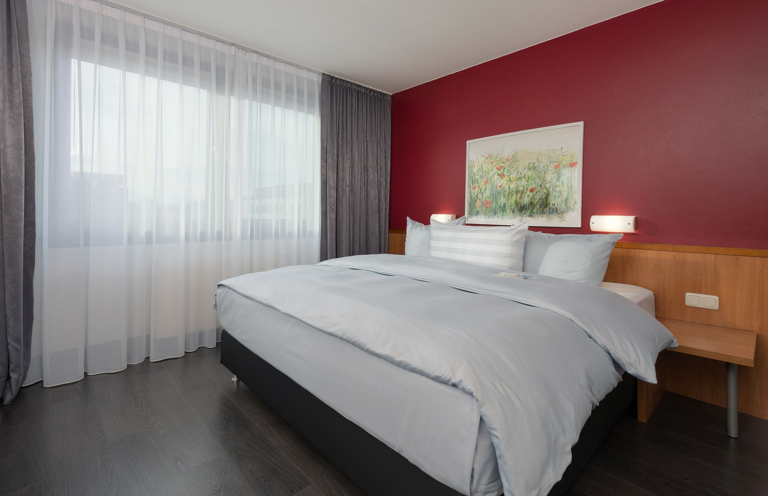 A modern hotel room with a large bed, a wooden nightstand, a painting on the wall, and sheer curtains on the window.