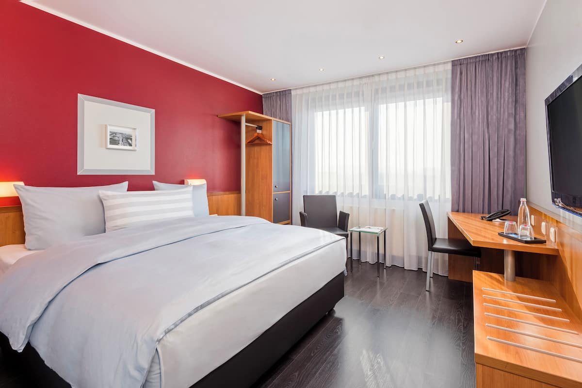 A hotel room with a bed, a desk, a chair, a TV, and a window with curtains.