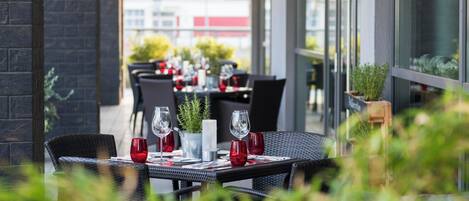 An outdoor dining area with tables set for two, featuring red glassware and a potted plant.