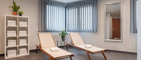 A modern spa room with two wooden lounge chairs, a white shelving unit with towels, a small table with a plant, and a large window with vertical blinds.