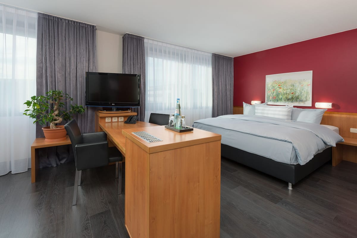 A hotel room with a bed, a desk with a TV, a potted plant, and a painting on the wall.