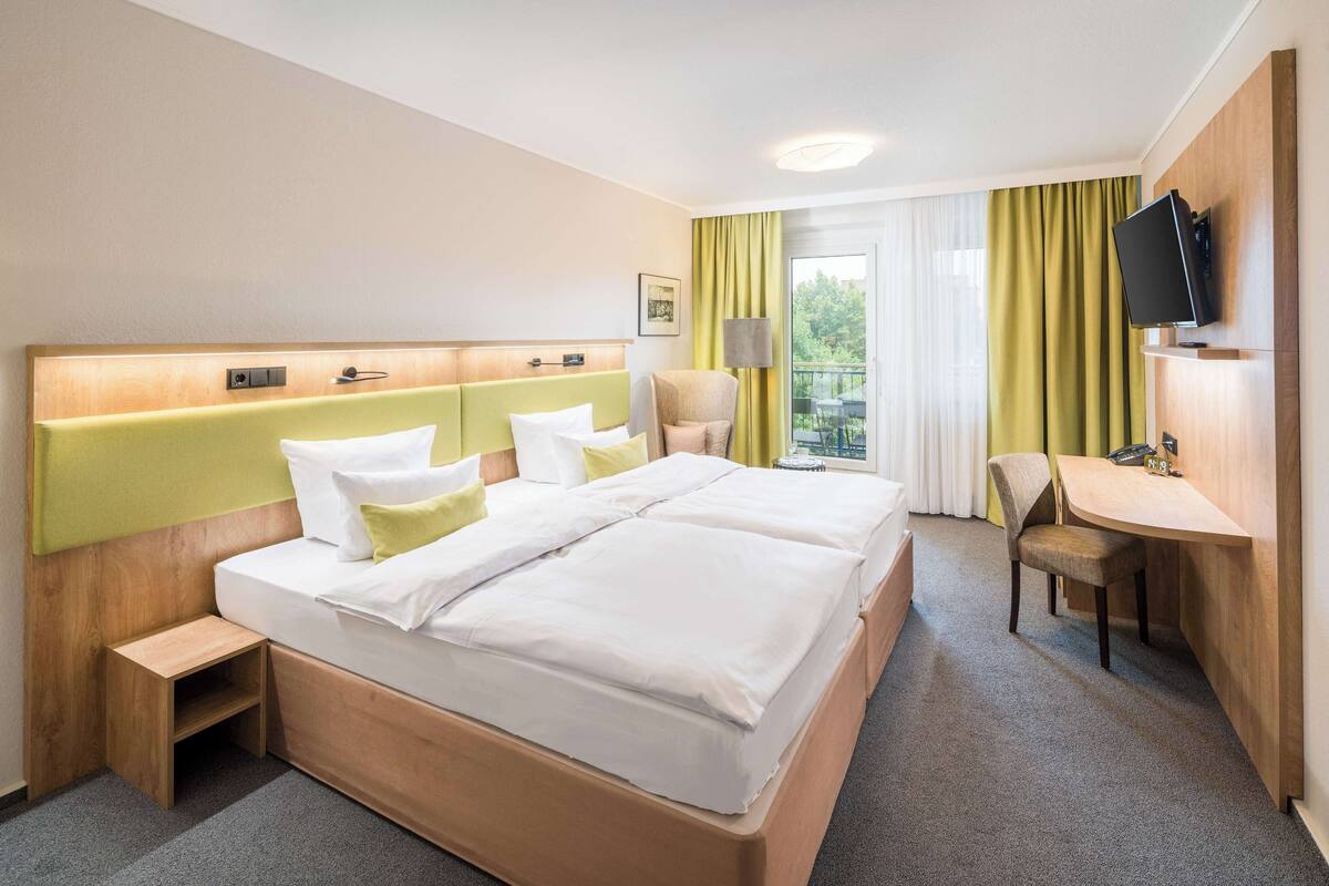 A hotel room with a double bed, a small desk, a chair, a television, and a window with curtains.