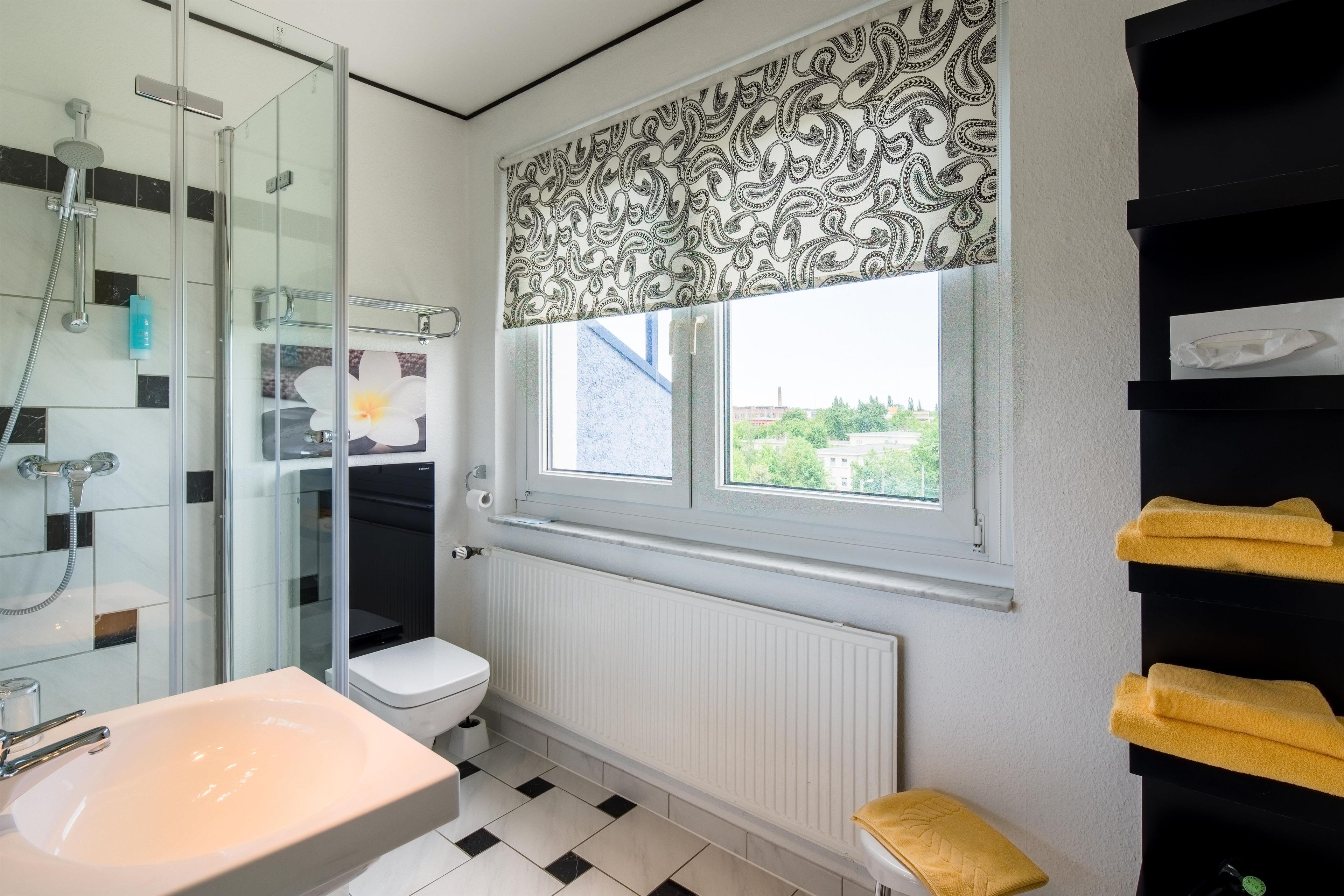 Ein Badezimmer mit Duschkabine aus Glas, Toilette, Waschbecken und Fenster mit blickdichter Jalousie.