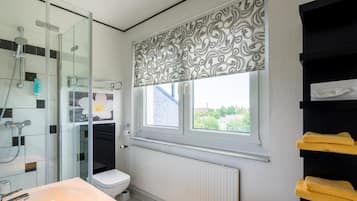Ein Badezimmer mit Duschkabine aus Glas, Toilette, Waschbecken und Fenster mit blickdichter Jalousie.