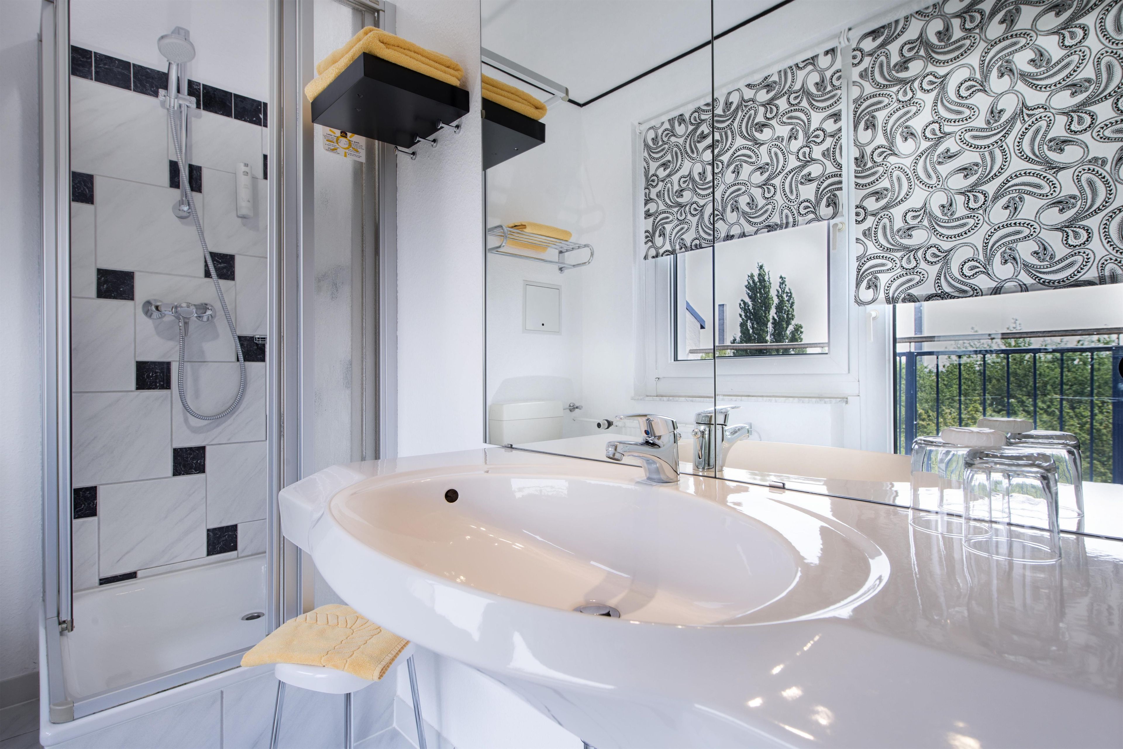 Ein modernes Badezimmer mit einem großen Waschbecken, einer Dusche mit Glastür und einem Spiegel mit dekorativem Muster.