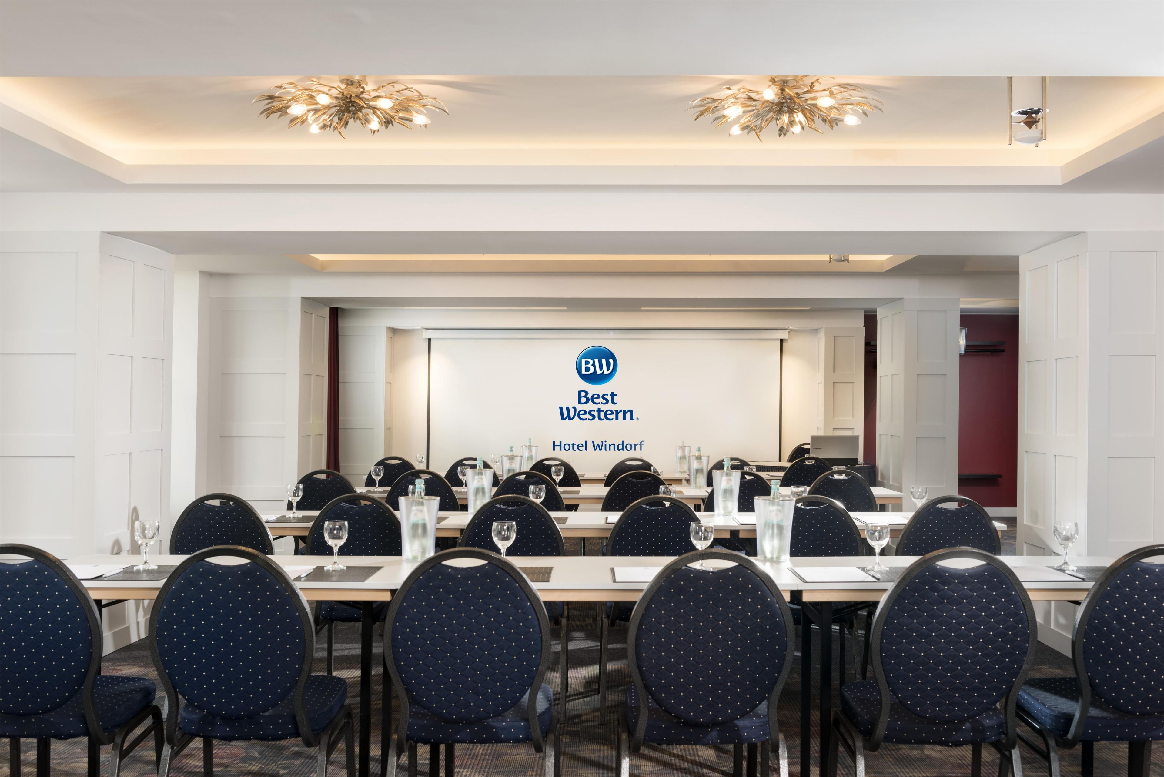 Ein Konferenzraum mit einem langen Tisch, Stühlen und einem Bildschirm, der das Logo des Best Western Hotel Windorf zeigt.