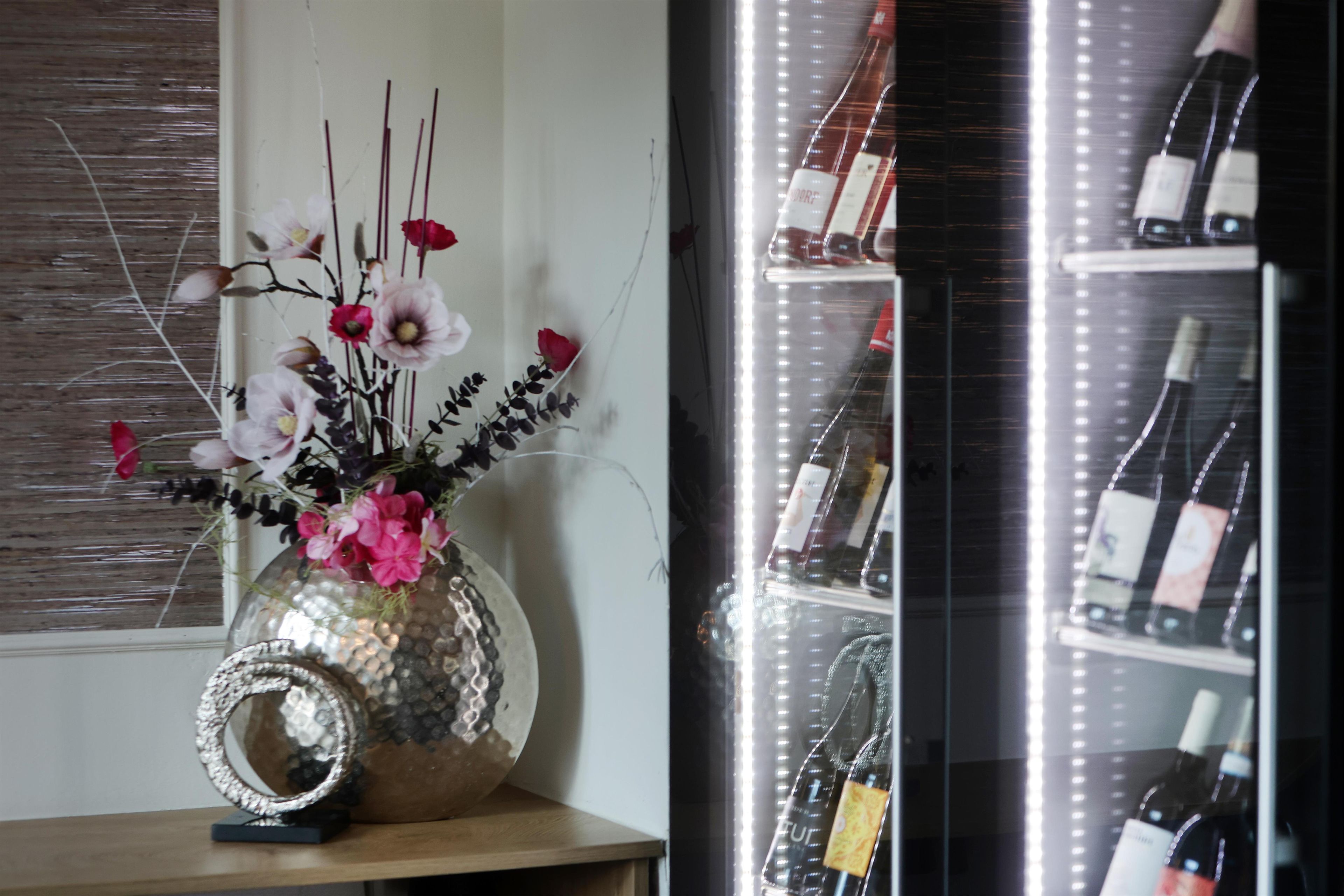 Eine dekorative Vase mit Blumen auf einer Holzoberfläche, ein Weinregal mit Flaschen und ein Fenster mit Jalousien.
