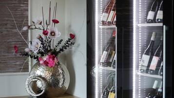 Eine dekorative Vase mit Blumen auf einer Holzoberfläche, ein Weinregal mit Flaschen und ein Fenster mit Jalousien.