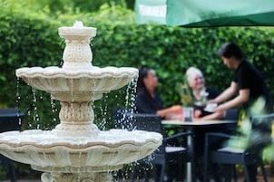 Una fuente de piedra de varios niveles con agua brotando del nivel superior, con un fondo de vegetación y un área de comedor con personas sentadas en mesas.