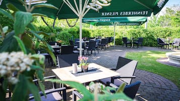 Un espace extérieur aménagé avec des tables et des chaises, protégé par un parasol vert orné de motifs, et entouré de verdure.