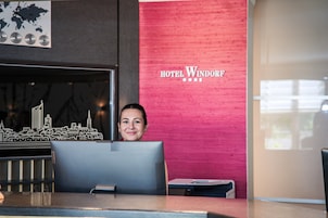 Un mostrador de recepción con un modelo de la ciudad, una pared roja con la señalización 'HOTEL WINDORF' y una ventana con luz natural.