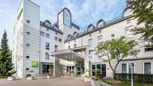 Ein modernes Hotel mit überdachter Eingangsparte, Außenbereich zum Sitzen und einem angelegten Garten.
