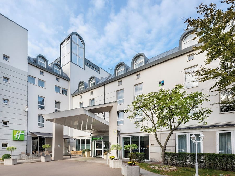 Ein modernes Hotel mit überdachter Eingangsparte, Außenbereich zum Sitzen und einem angelegten Garten.