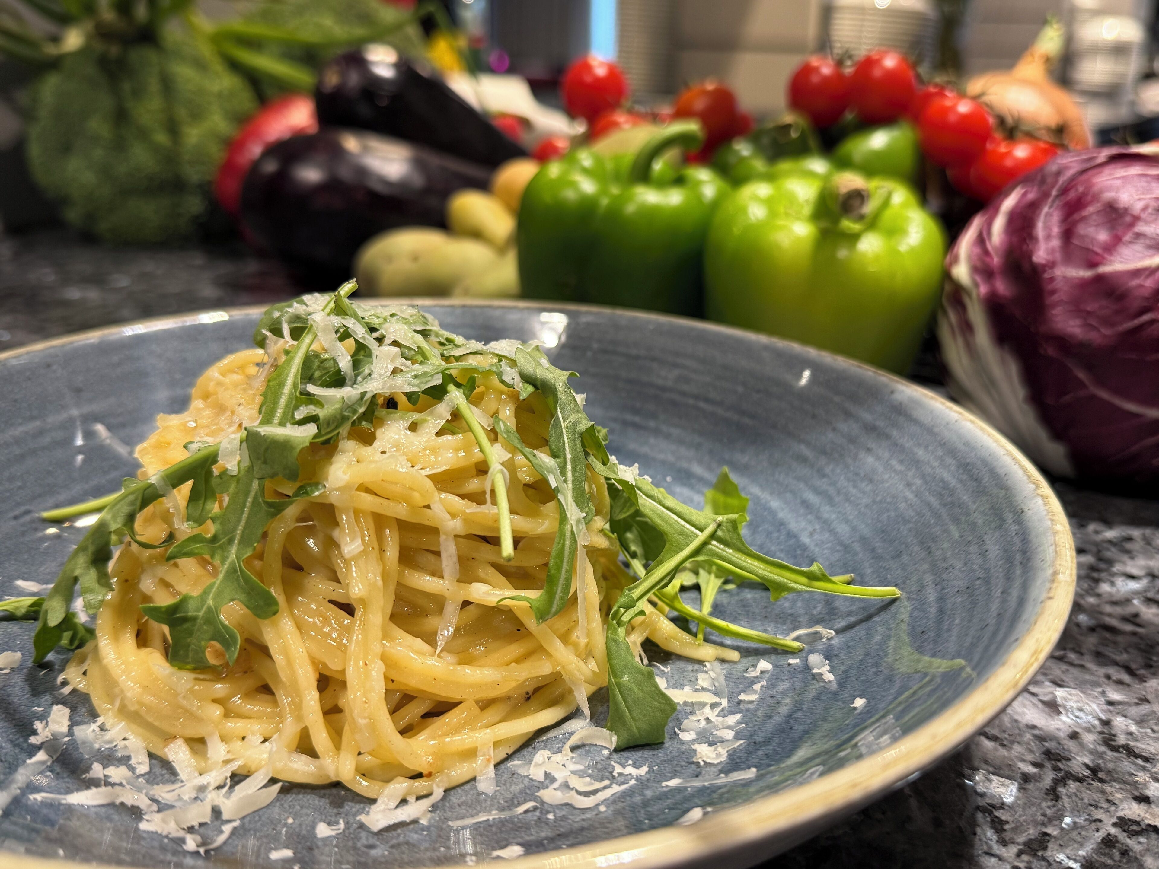A plate of spaghetti garnished with greens and cheese, with vegetables in the background.