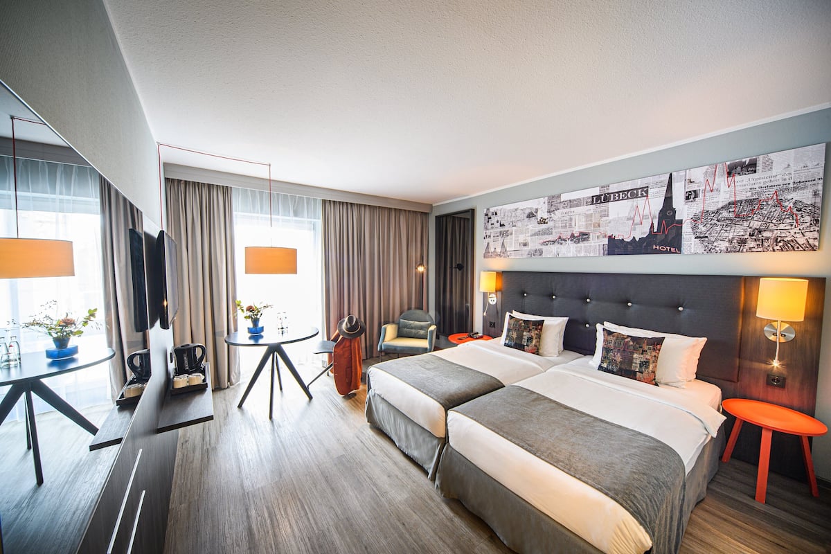 Habitación de hotel con una cama grande, un escritorio con una silla, una mesita con una lámpara y un ventanal con cortinas.