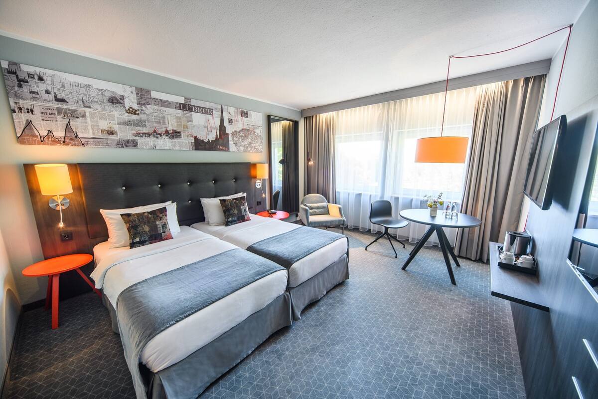 Quarto de hotel com duas camas, uma escrivaninha, uma cadeira, uma televisão e um mural urbano na parede.