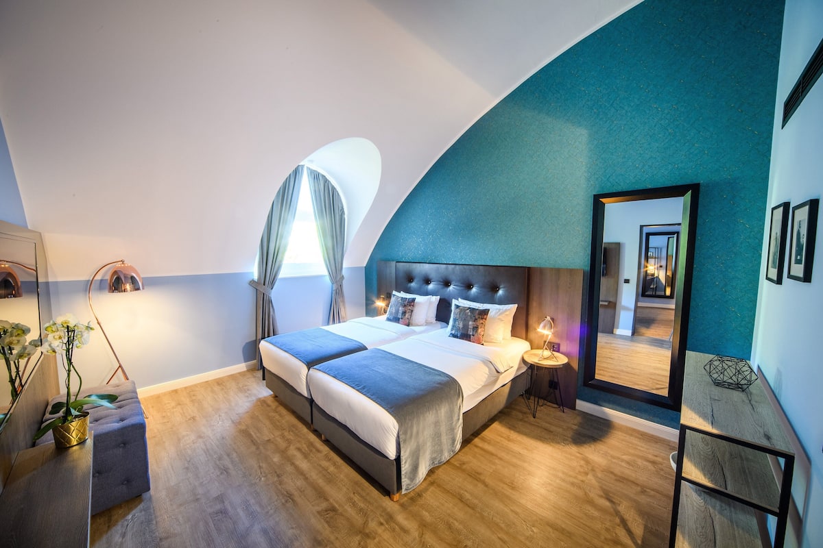 Quarto moderno com uma cama grande, mesa de cabeceira, espelho e vista para o corredor.