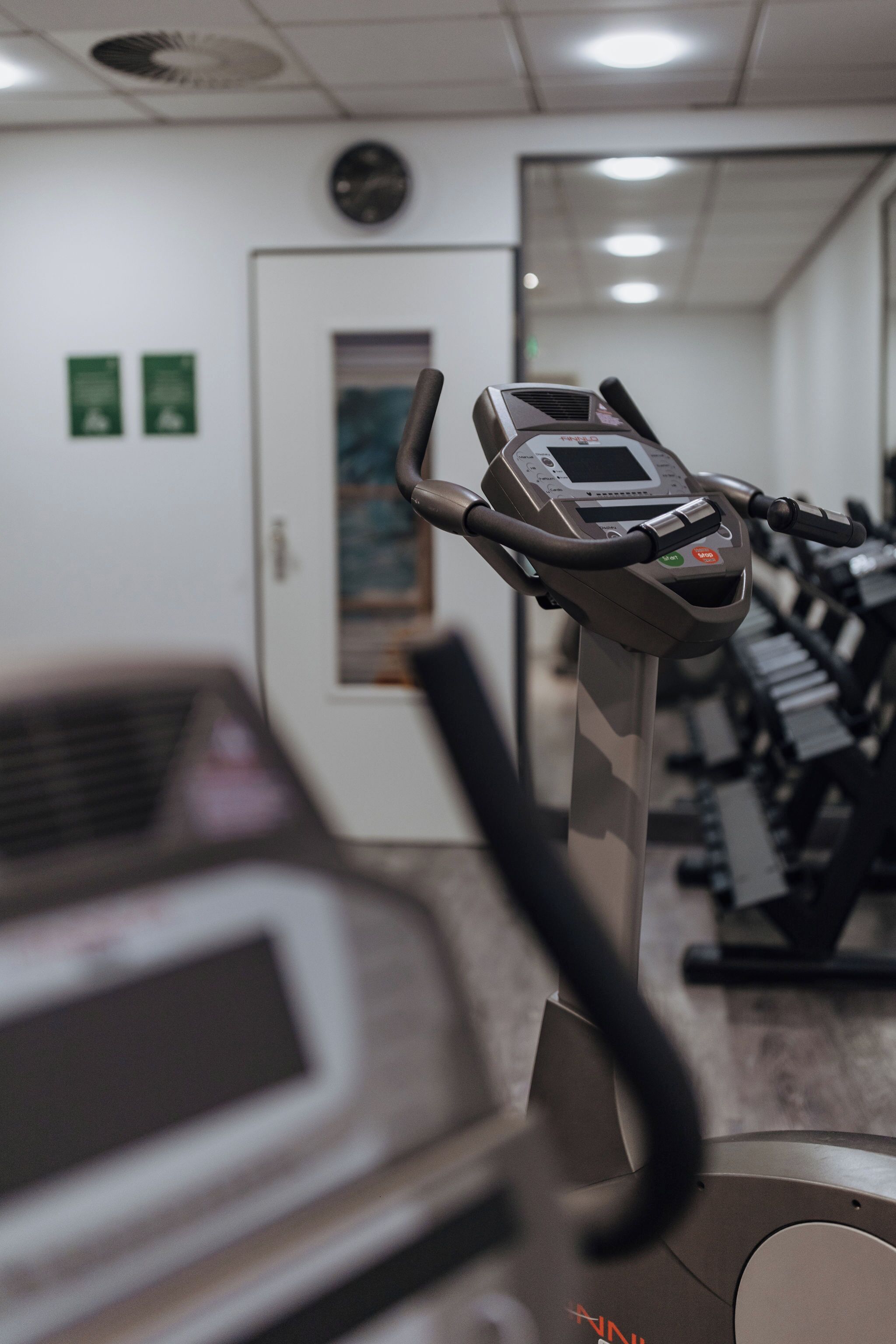 Una cinta de correr en un gimnasio, con un muro espejado y una puerta al fondo.
