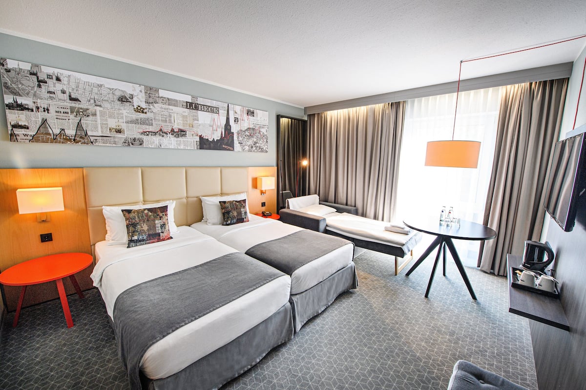 Quarto de hotel com duas camas, uma mesinha, um sofá, uma televisão e um mapa grande na parede.