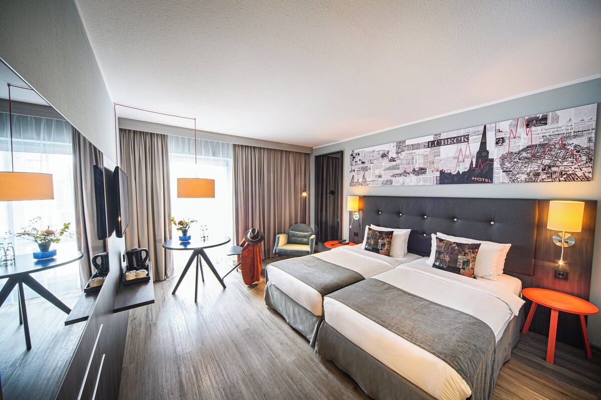 A hotel room with a large bed, a desk with a chair, a small table with a lamp, and a large window with curtains.