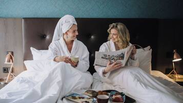 Two people in white robes sitting on a bed with a tray of food and drinks, reading a magazine.