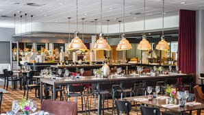 A spacious restaurant with multiple dining tables and chairs, overhead pendant lighting, and a bar area in the background. - Holiday Inn Luebeck by IHG (Lübeck)