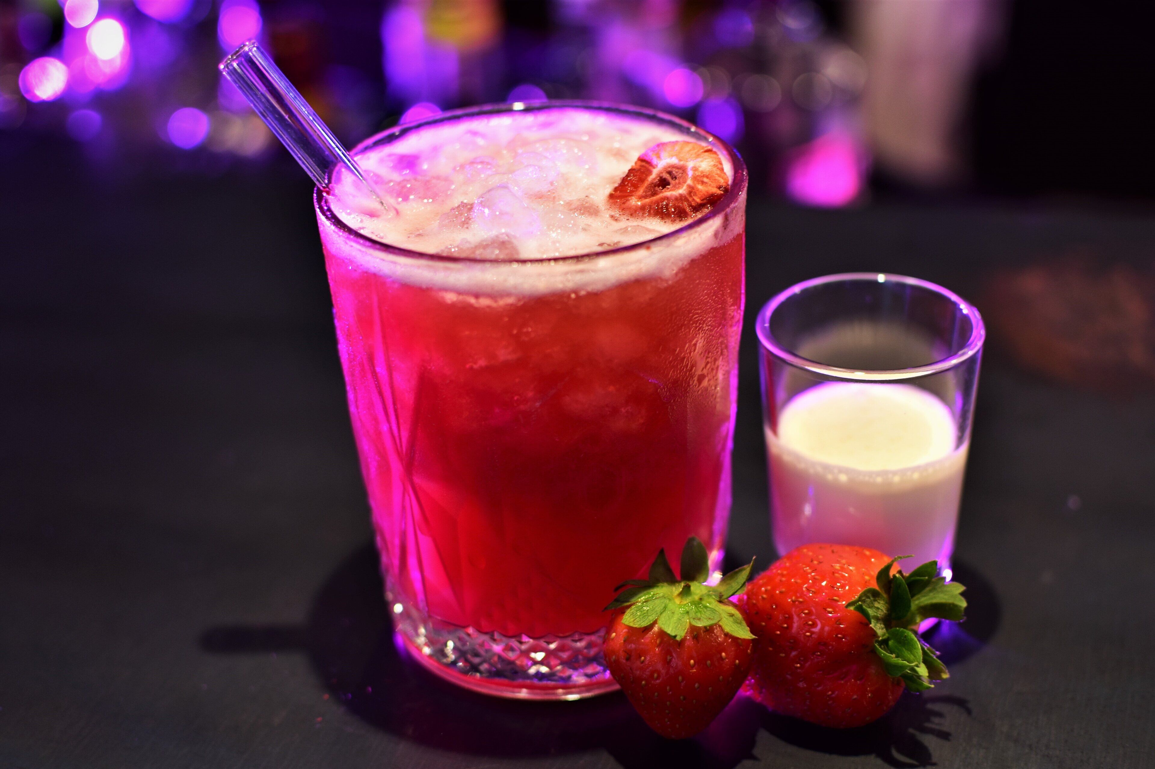 A cocktail glass with a strawberry garnish, a glass of milk, and two strawberries on a table.