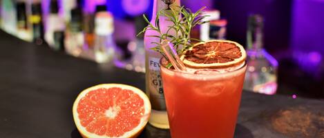 A glass of red beverage with a slice of citrus and a sprig of rosemary, accompanied by a half grapefruit and a bottle labeled "FEVER."