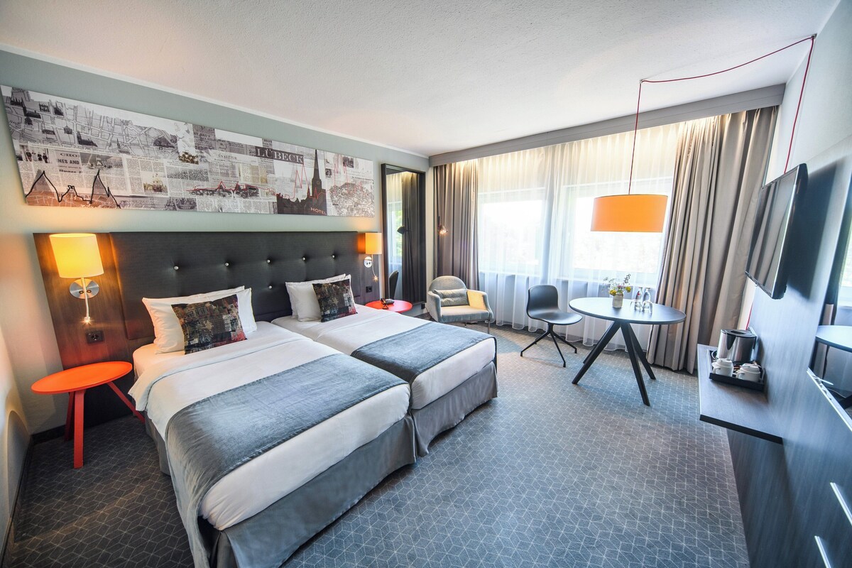 A hotel room with two beds, a desk, a chair, a TV, and a wall with a cityscape mural.