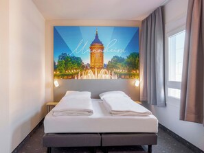 A hotel room with a bed, a large wall-mounted picture of a famous landmark, and a window with curtains.
