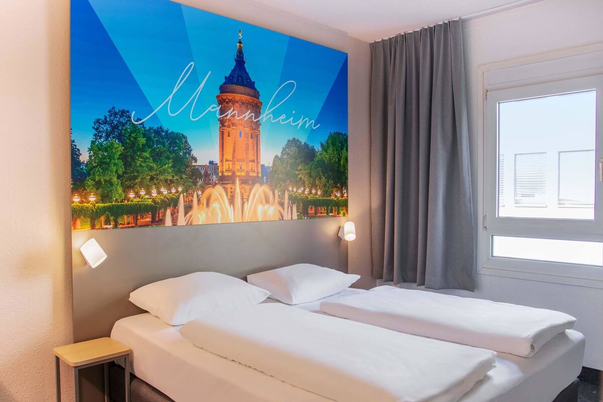 A hotel room with a bed, a nightstand, a wall-mounted lamp, and a window with curtains.