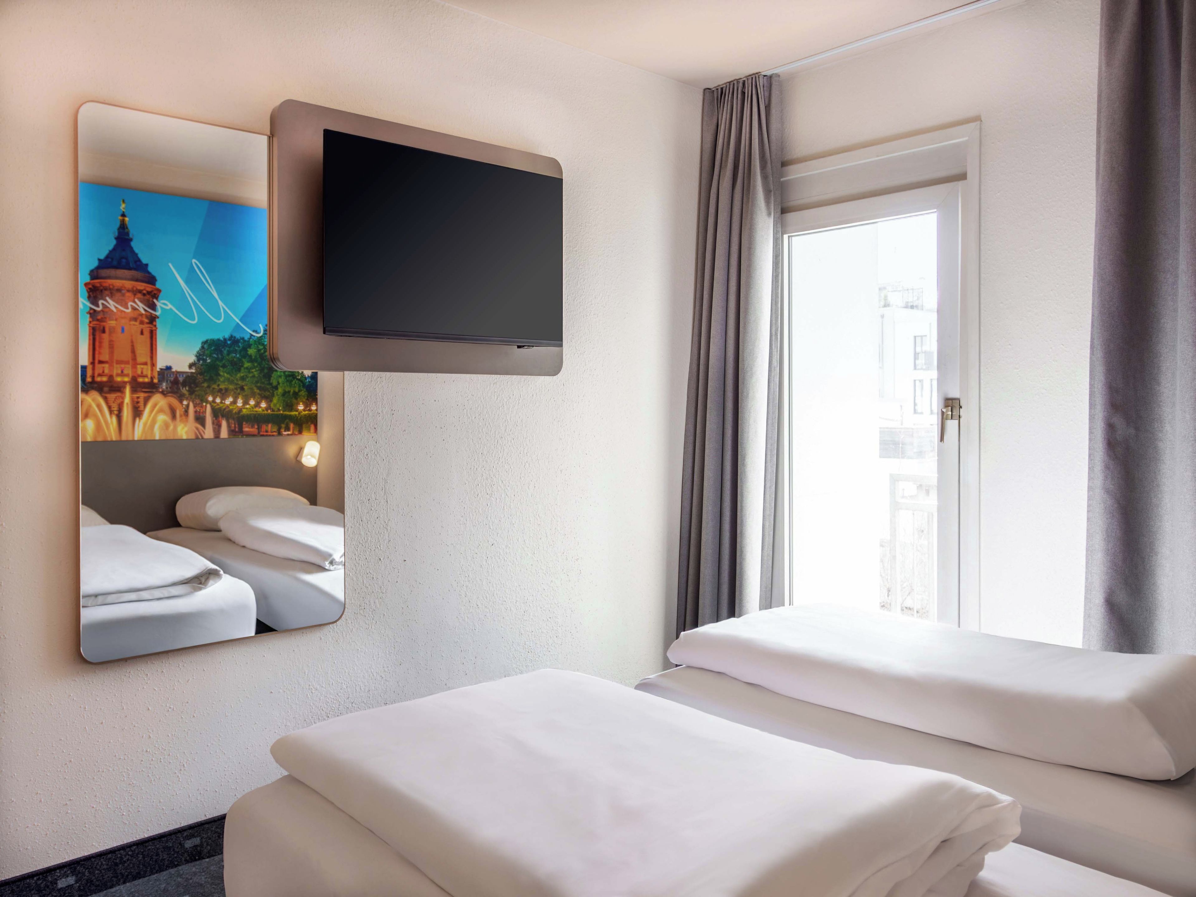 A hotel room with a flat-screen TV mounted on the wall, a bed with white bedding, a window with curtains, and a painting of a bridge.